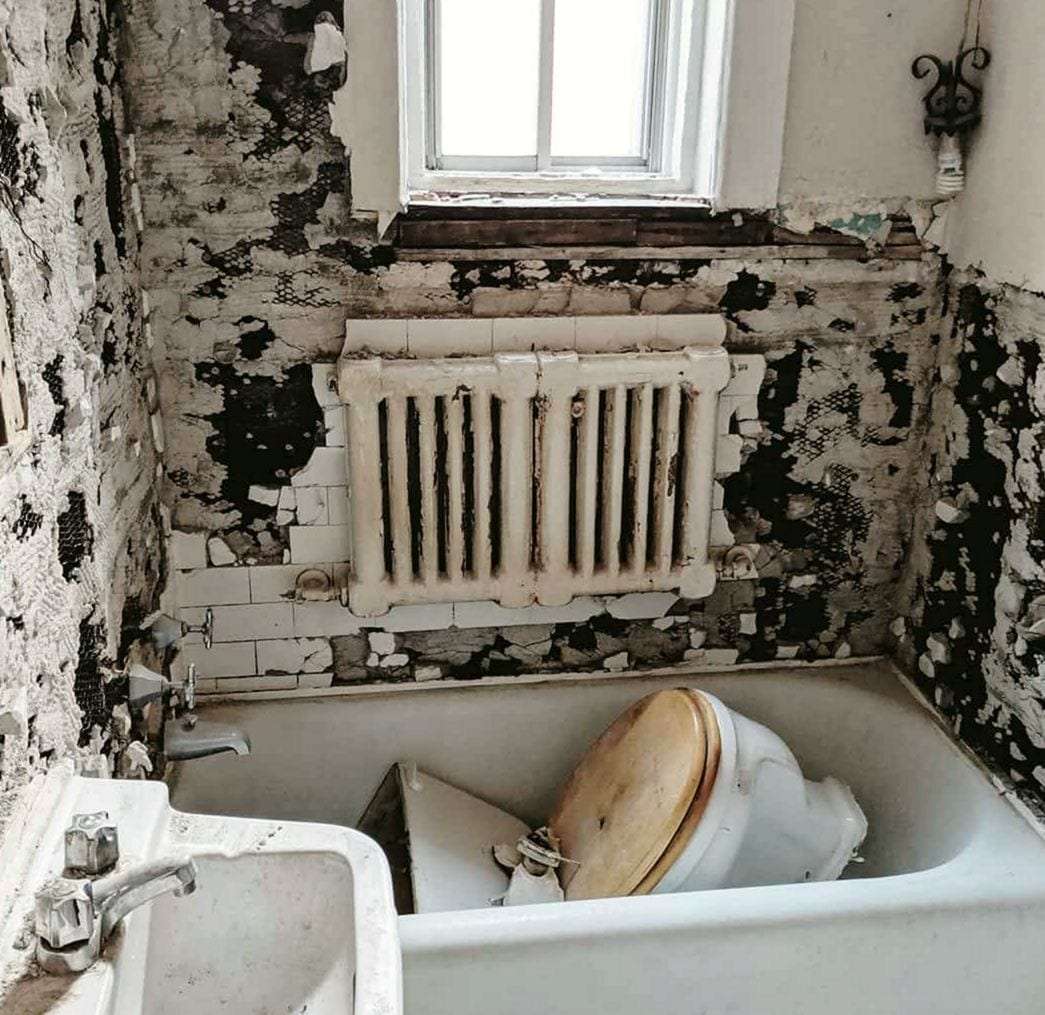 Old bathroom stripped to the plaster, the toilet set in the tub beside a wall sink and radiator