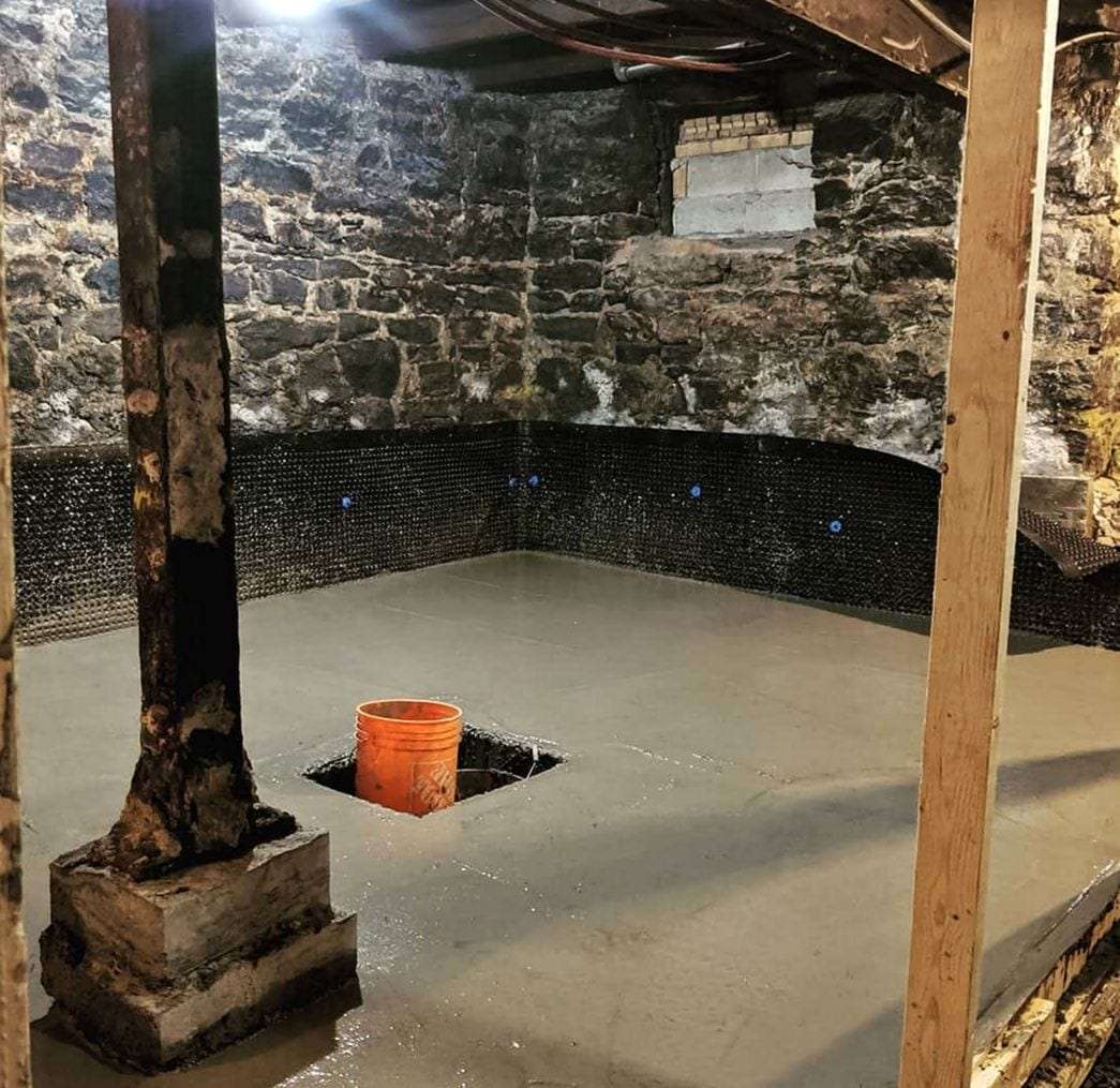 Stone-walled basement with a fresh concrete floor, drainage membrane along the walls and an open sump pit