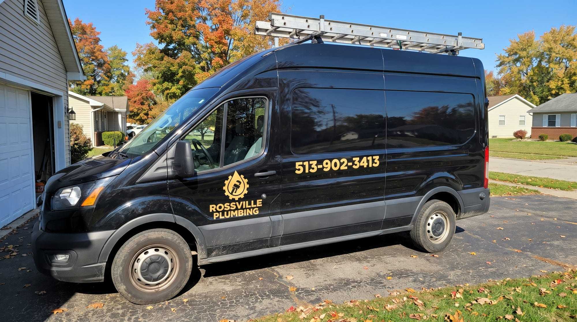 Black Rossville Plumbing cargo van with gold lettering and a ladder rack, parked in a driveway beside a garage under autumn maples