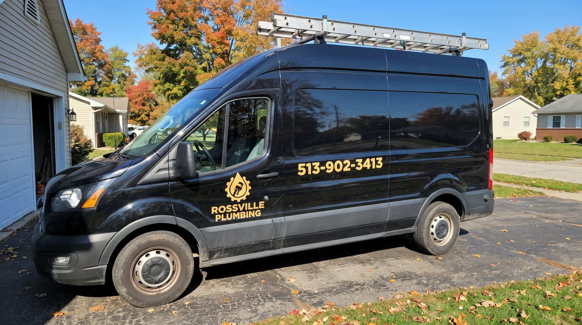 Black Rossville Plumbing cargo van with gold lettering and a ladder rack, parked in a driveway beside a garage under autumn maples